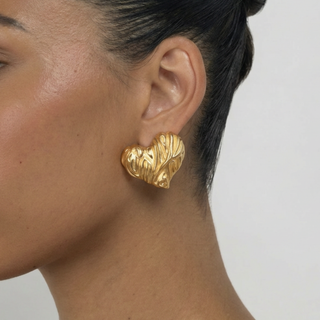 Gold heart-shaped earring worn by a person on a neutral background