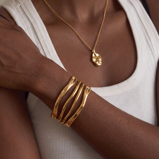 A close-up of a person wearing a gold cuff bracelet with wavy parallel bands on their wrist, along with a matching gold necklace with a textured pendant, over a white ribbed tank top.